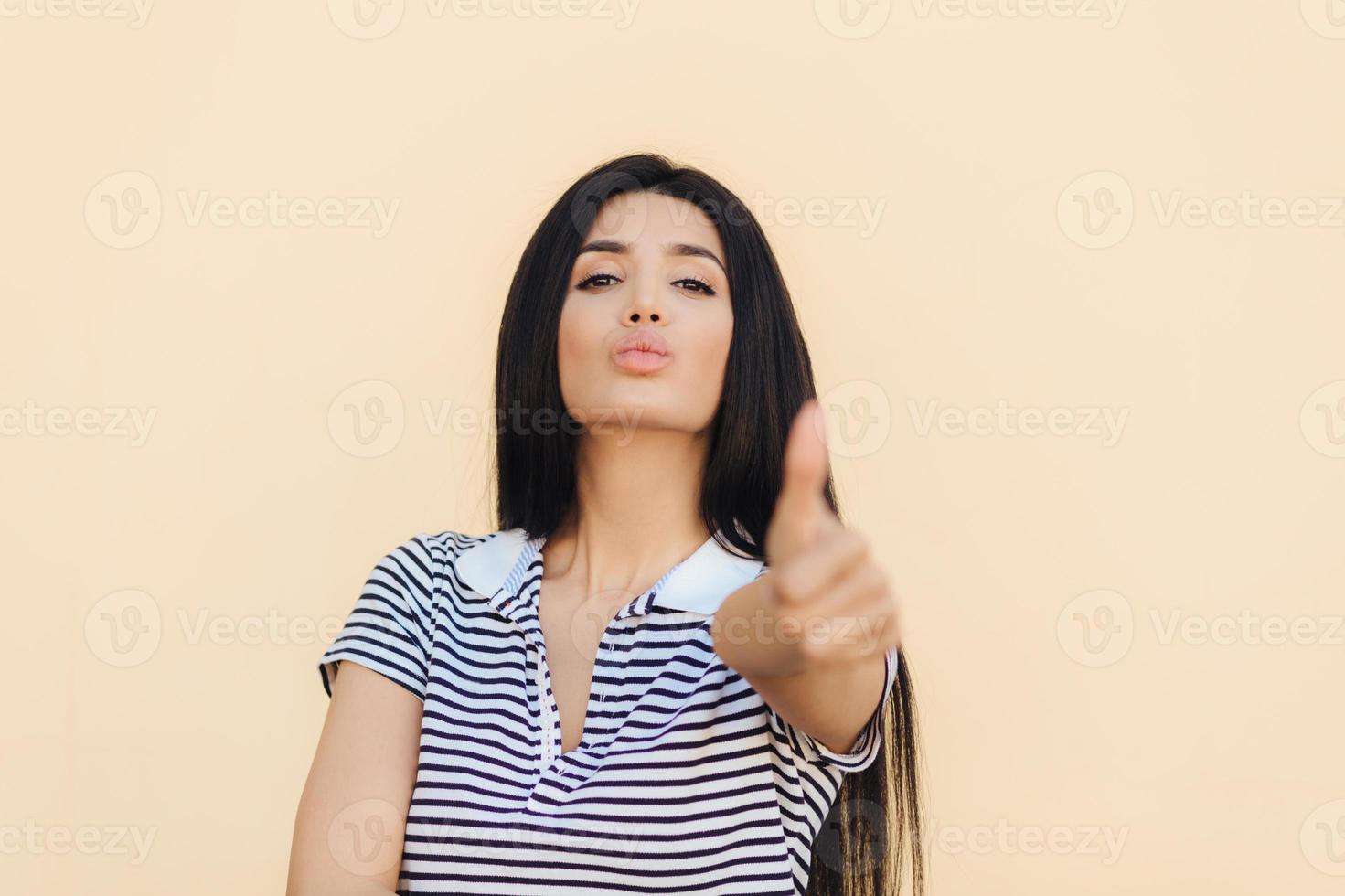 Confident pleased young woman with self assured expression, keeps thumb raised, shows approval gesture, has healthy skin, lips round, isolated over beige background. People and body language photo