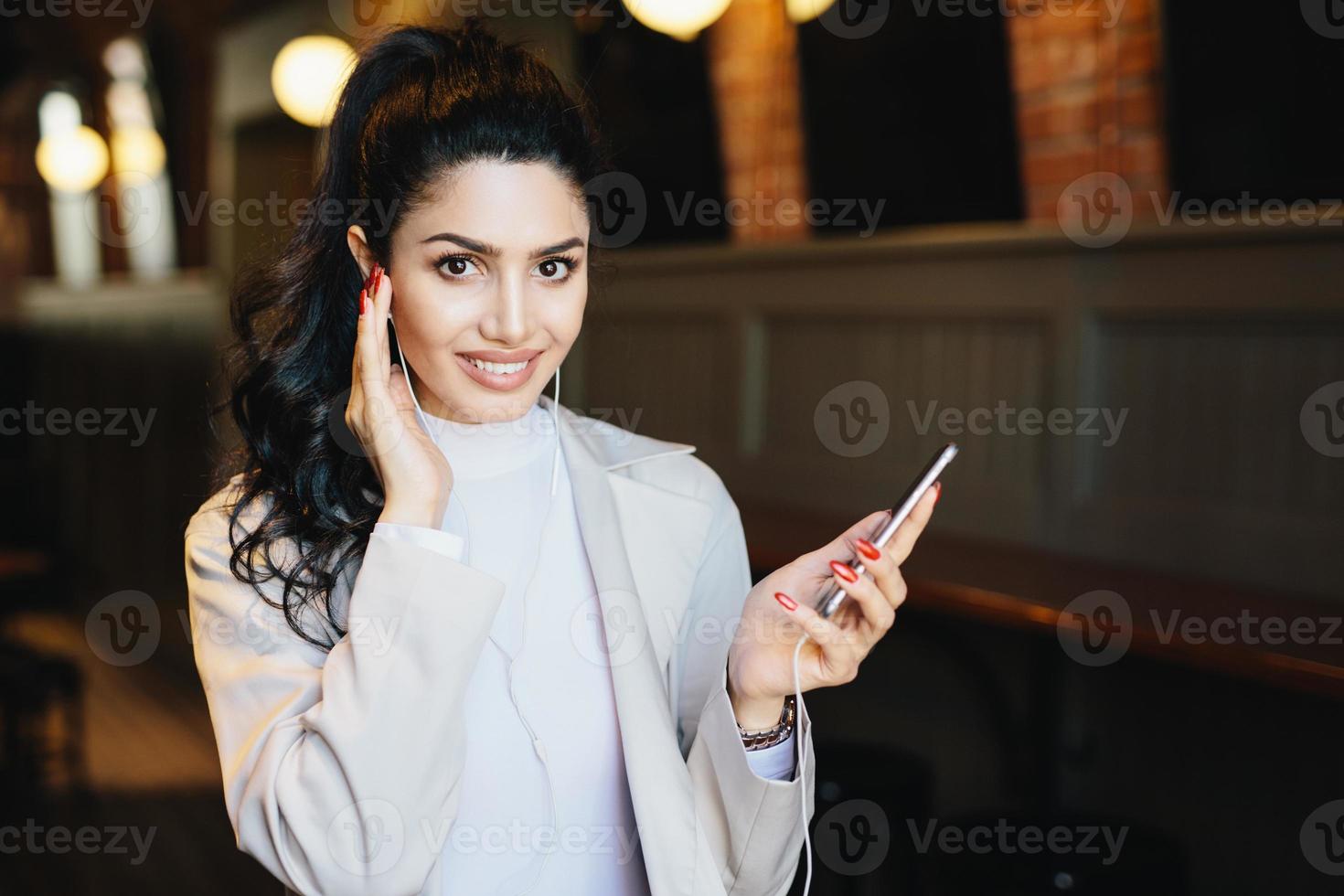 Cute woman with dark pony tail having long eyelashes and expressive eyes sitting at cafe listening to music online enjoying high-speed internet connection. People, beauty and technology concept photo