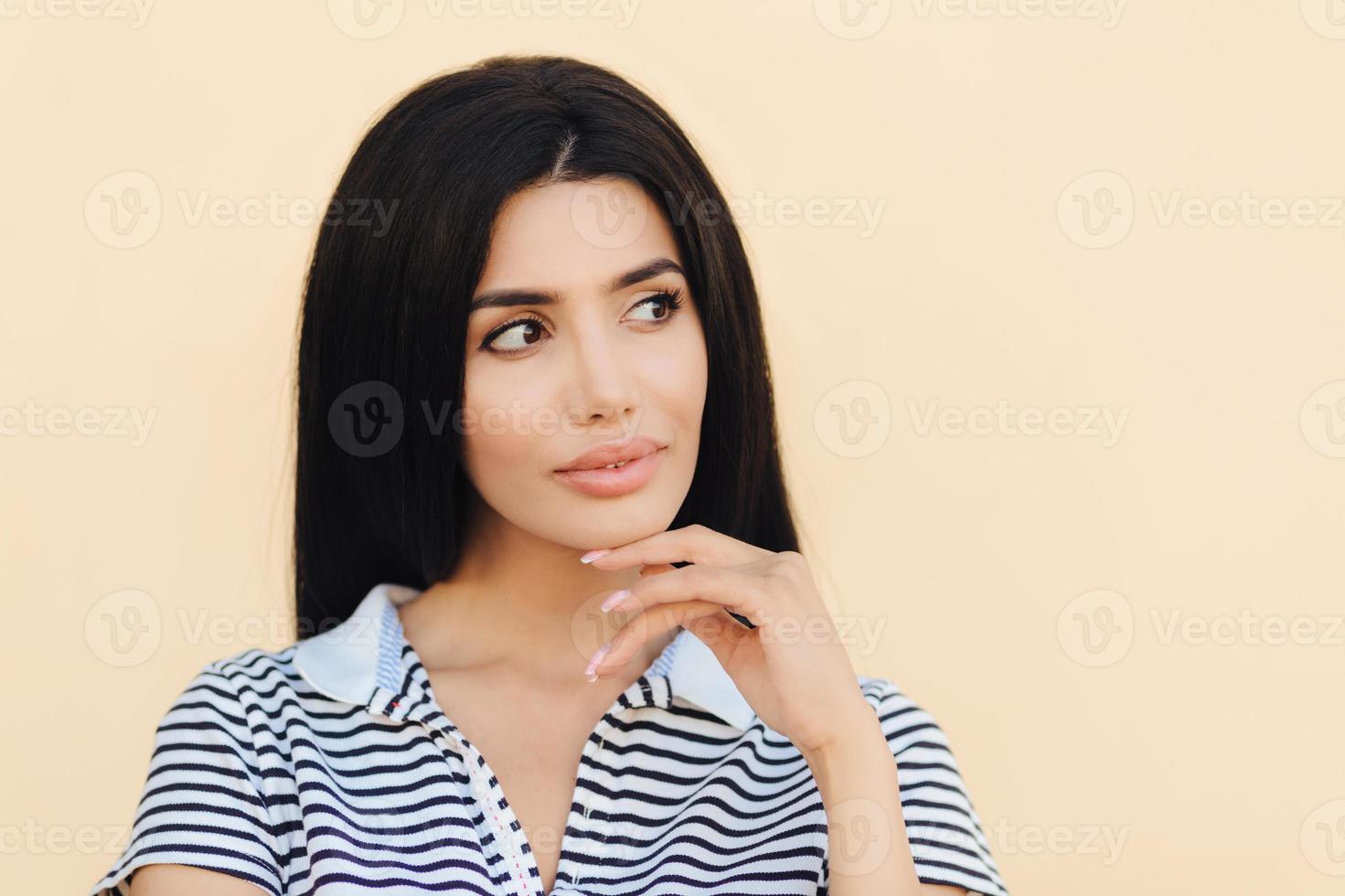 Close up portrait of attractive brunette female with healthy skin, looks pensively aside, keeps hand under chin, dreams about something pleasant, isolated over light pink background. Sensual cute lady photo