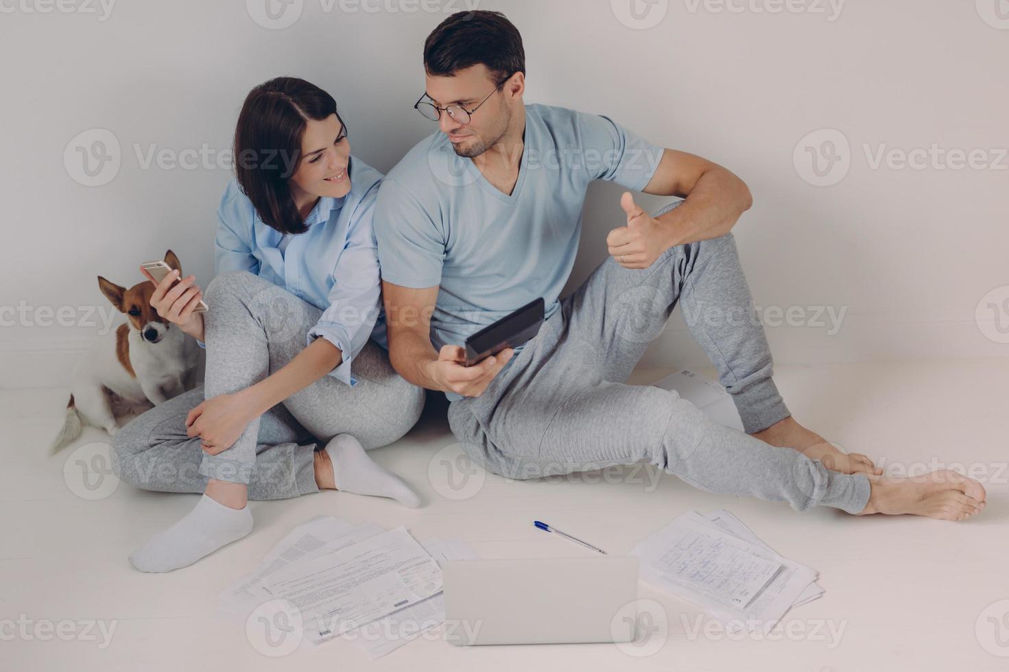 Photo of satisfied man wears spectacles and casual clothes, shows figures on calculator, demonstartes okay gesture, rejoices high profits, works together with female colleague, sit on floor.