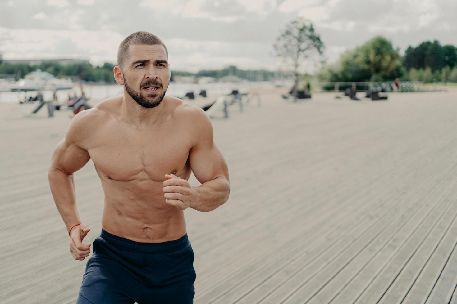 Outdoor shot of motivated shirtless muscular Caucasian man prepares for running marathon, goes jogging early in morning regularly, concentrated somewhere into distance. Sport and success concept photo