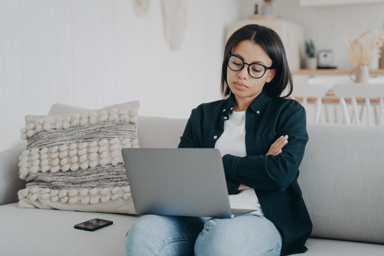 una mujer de negocios desconcertada con una laptop considera la solución del problema, brazos cruzados, sentada en el sofá en casa foto