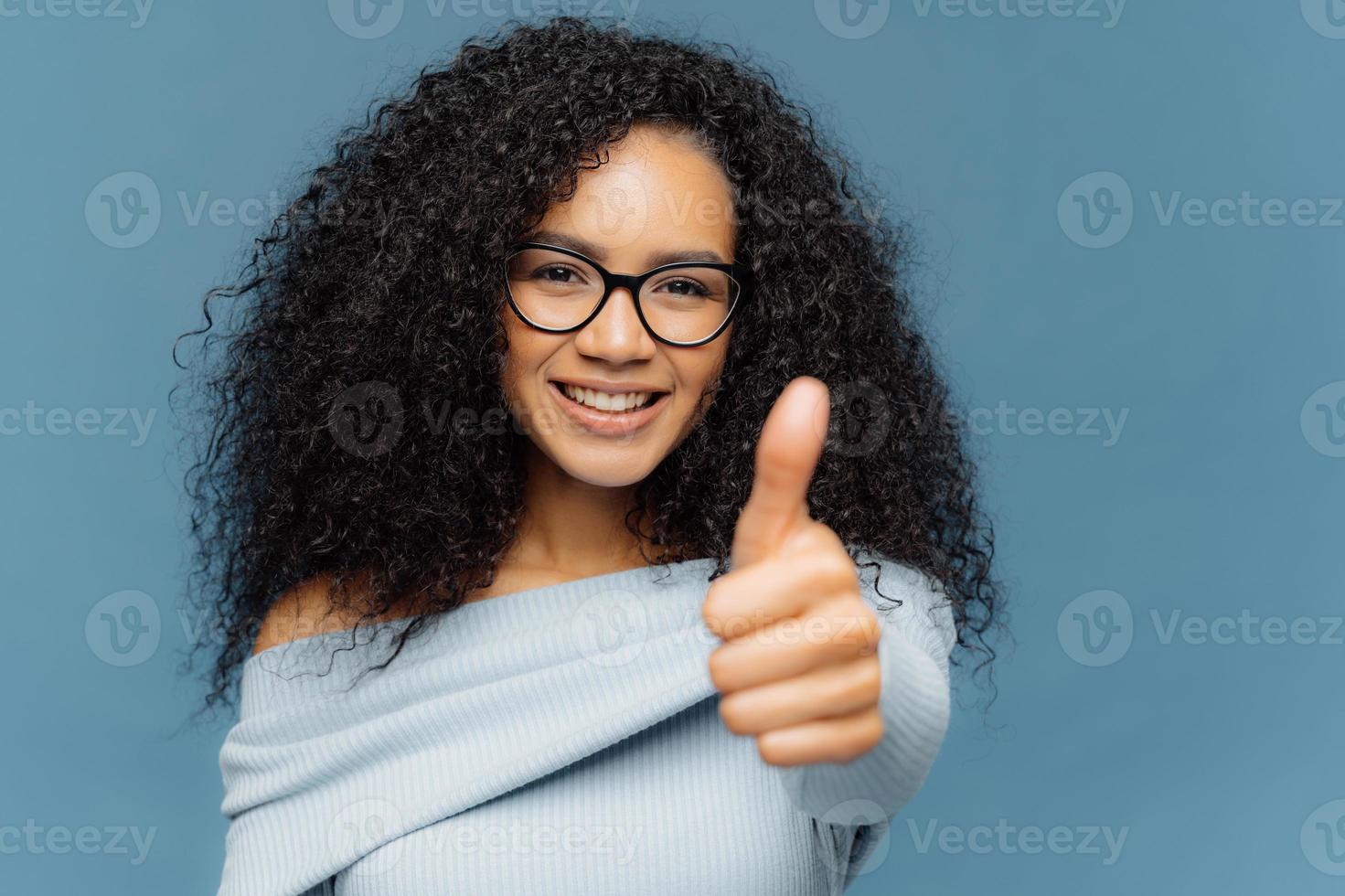 Portrait of dark skinned female has thumb up, shows agreement or approval, has smile on face, wears spectacles and jumper, gestures indoor against blue background. Body language and okay concept photo