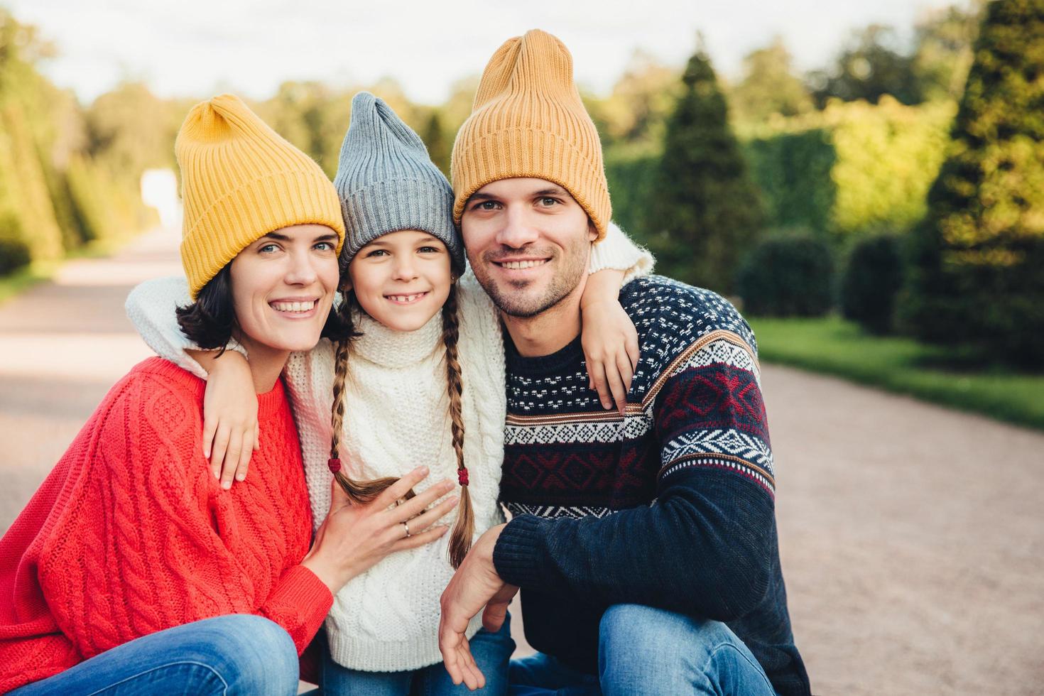 retrato horizontal de una familia afectuosa y amistosa que se abraza, usa gorras y suéteres de punto, caminan juntos, tienen sonrisas agradables en las caras. pareja casada e hija pequeña disfrutan del tiempo libre foto