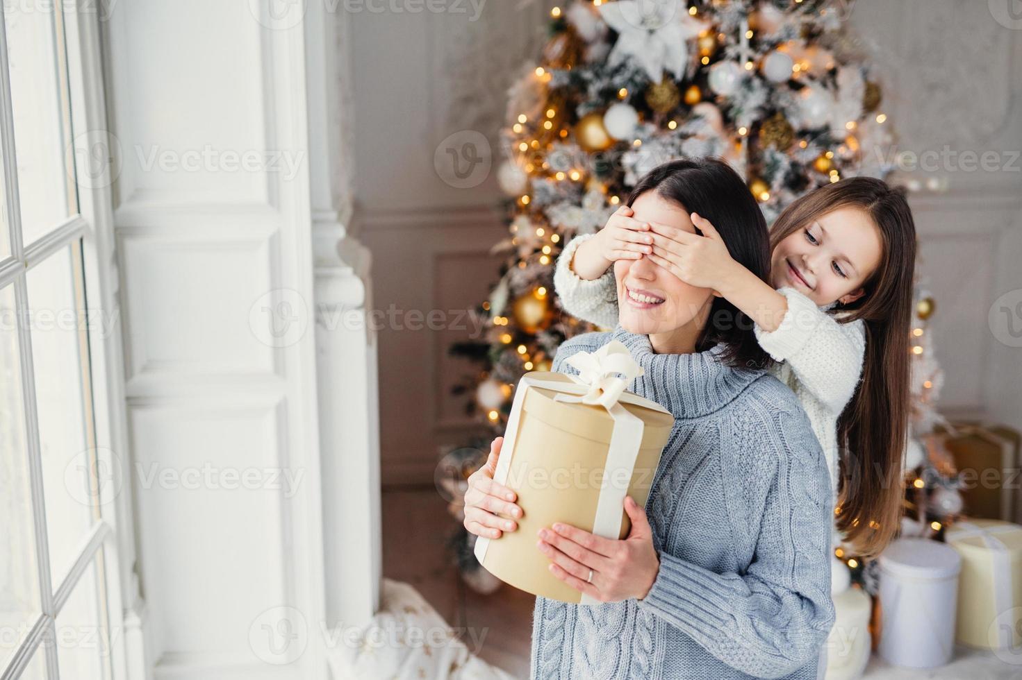 Portrait of little girl closes mothers eyes, congratulates her with New Year or Christmas, stand near window in living room, have real miracle and feeling of holiday. Winter, celebration, season photo