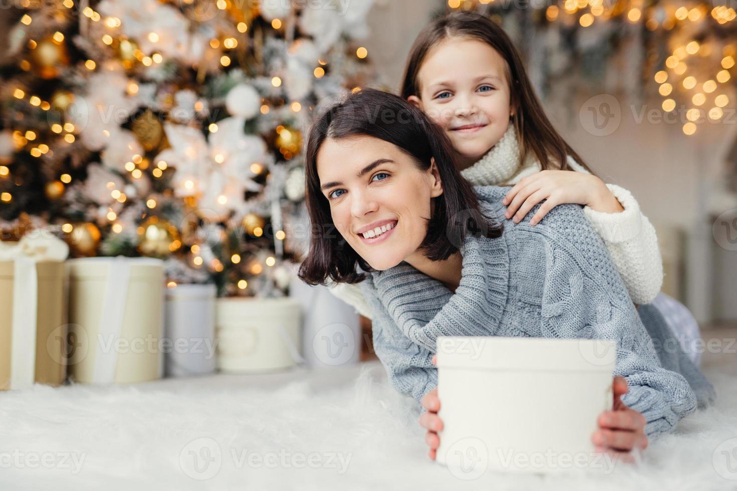 Brunette smiling female adult and her beautiful adorable small kid in white sweater being in living room, hold present, going to congratulate father and husband with coming New Year or Christmas photo