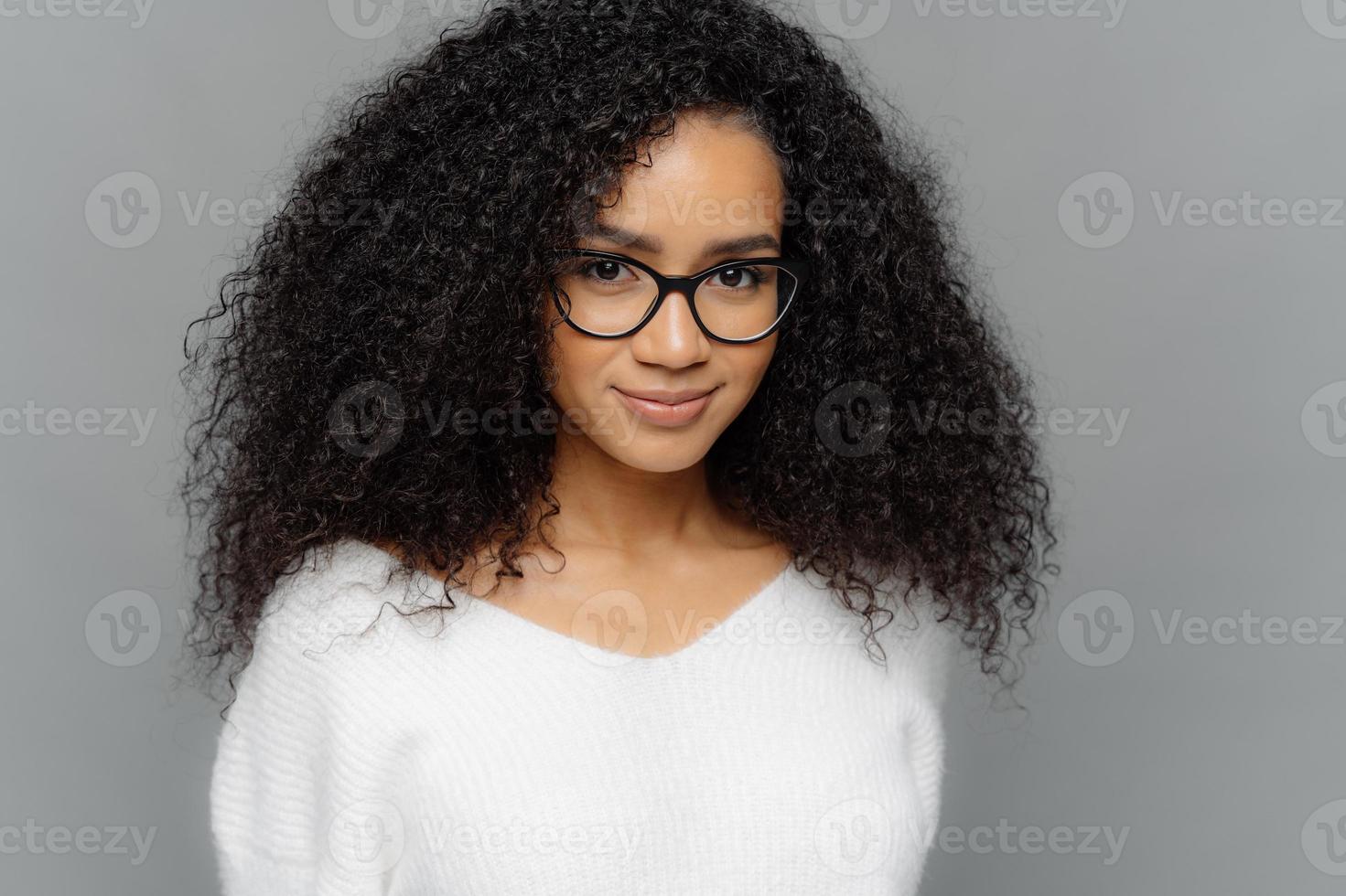 Close up shot of dark skinned female has satisfied expression, bushy curly hair, wears spectacles and white jumper, looks straightly at camera, models over grey background. Natural beauty concept photo