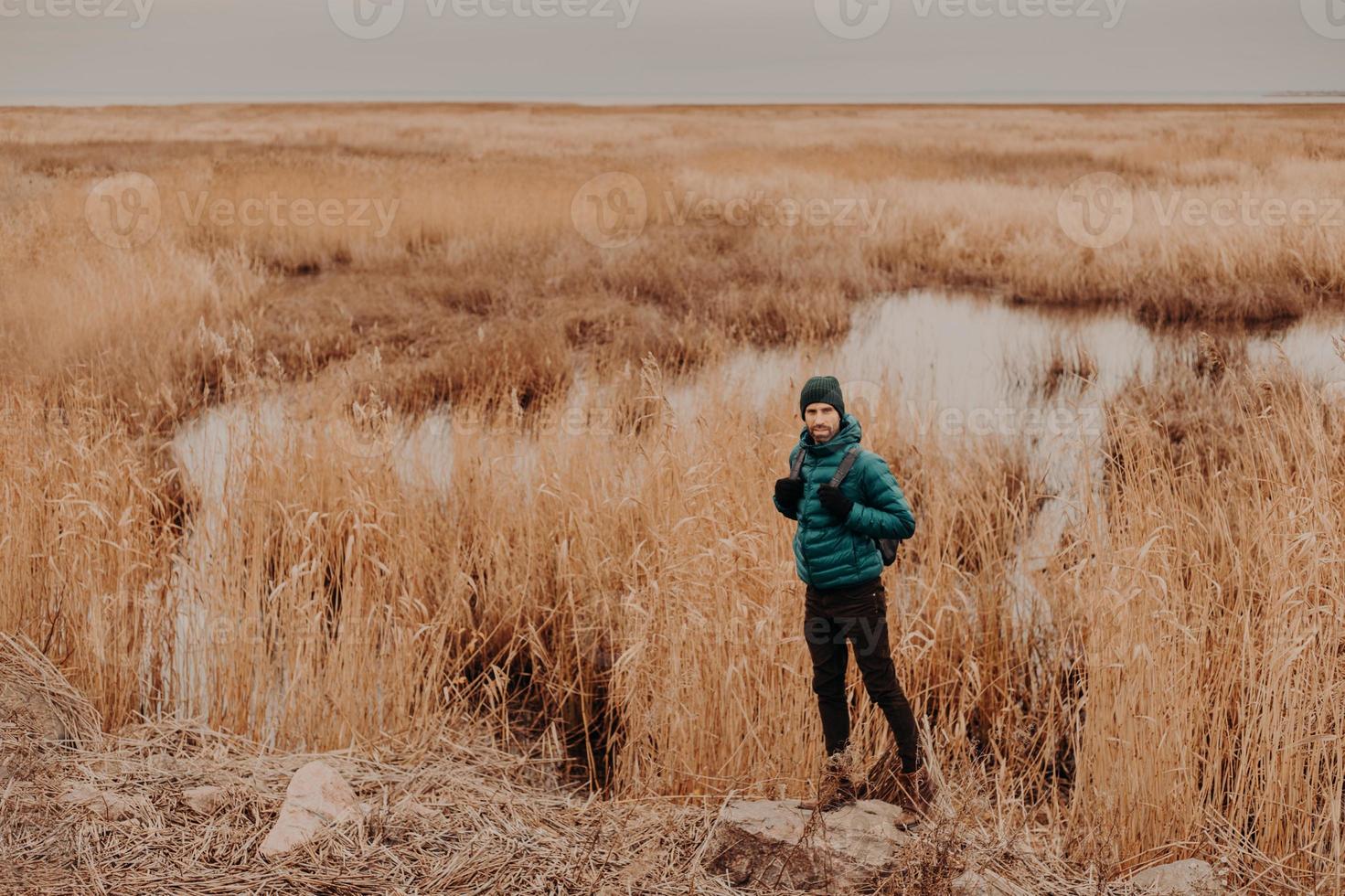 Photo of active male dressed in fashionable clothes, stands on stone against near small lake and yellow autumn field, carries backpack, copy space aside for your advertising content or information
