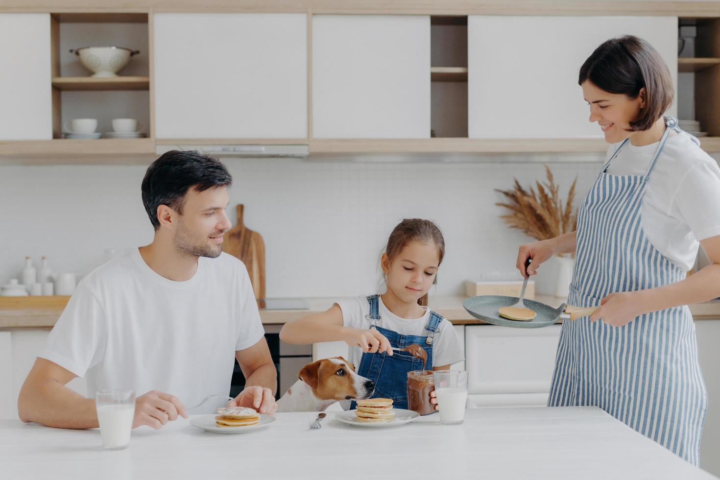 Family have breakfast at kitchen in morning. Happy girl puts melted chocolate on tasty fried pancake, poses at table with father, dog, mother stands near, wears apron, holds pan, busy cooking photo