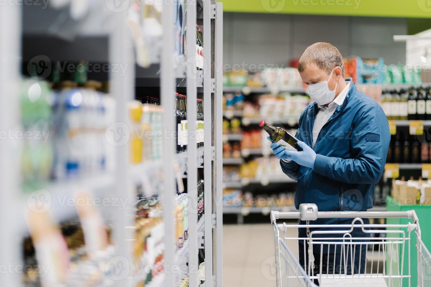 Photo of man in protective medical mask and rubber gloves to prevent coronavirus, poses in supermarket, holds glass bottle of alcoholic drink, reads label, makes shopping during quarantine time