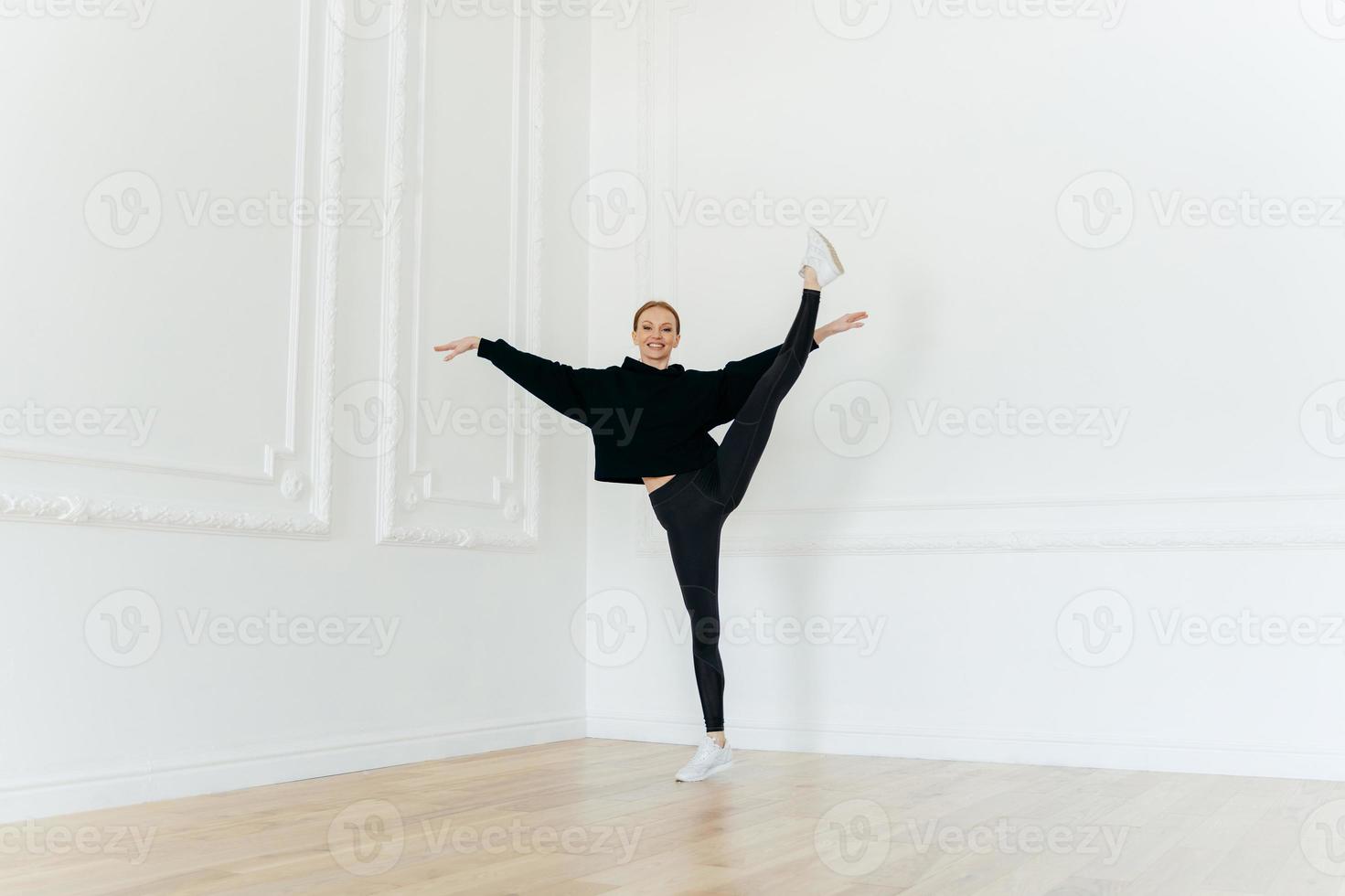 Gorgeous flexible girl stands on one leg, does stretching exercises, wears black sportswear, balances indoor, has satisfied look. Sporty woman practices yoga. People, wellness and fitness concept photo