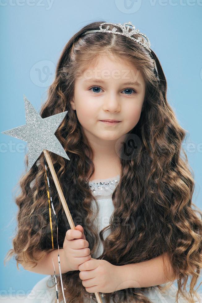 Small kid angel has long crisp hair, blue eyes, holds magic wand in form of star, poses over blue background, has appealing appearance. Little princess waits for miracle, believes in fairy tale photo