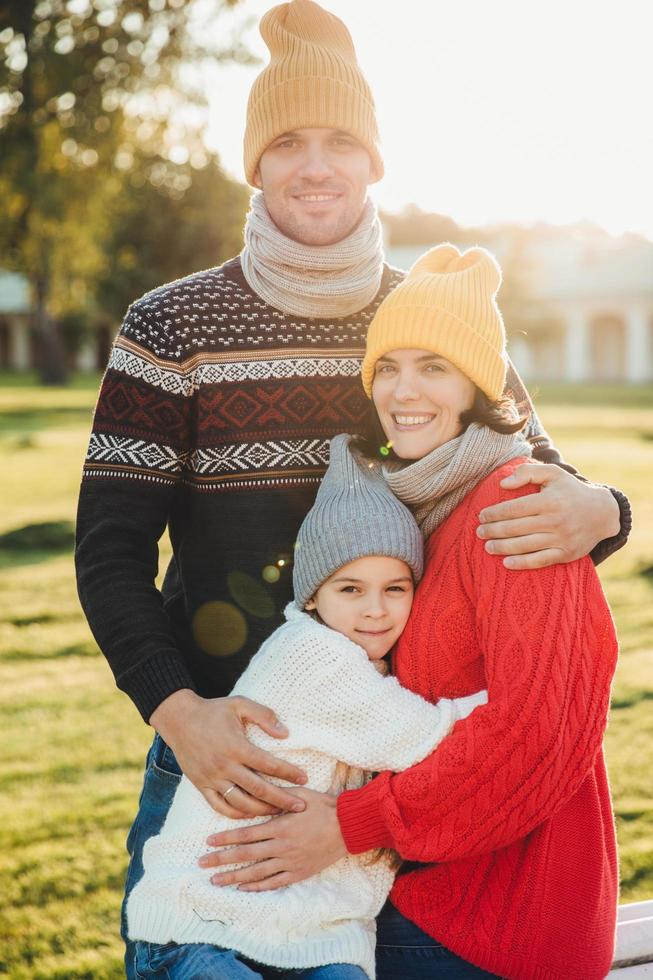 retrato vertical de una pareja familiar amistosa que se une, se abraza, tiene buenas relaciones, disfruta del clima soleado. hombre guapo sin afeitar abraza a su esposa e hija, posa al aire libre foto