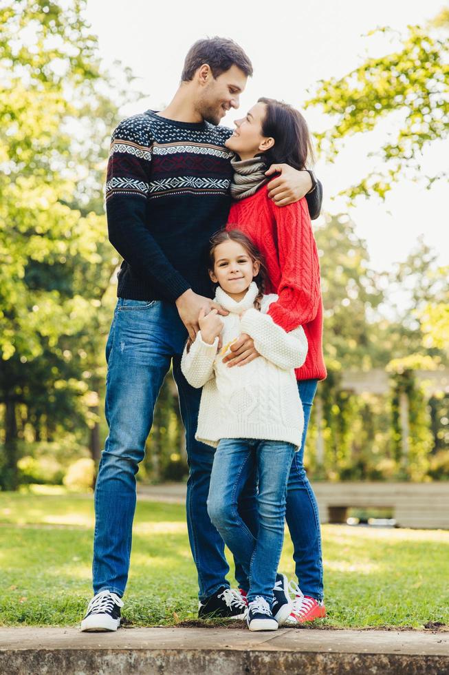 Young happy family of three stand together outdoors, embrace, have good relationships. Couple in love look at each other, hug their little daughter. Happiness and harmony in family life. photo