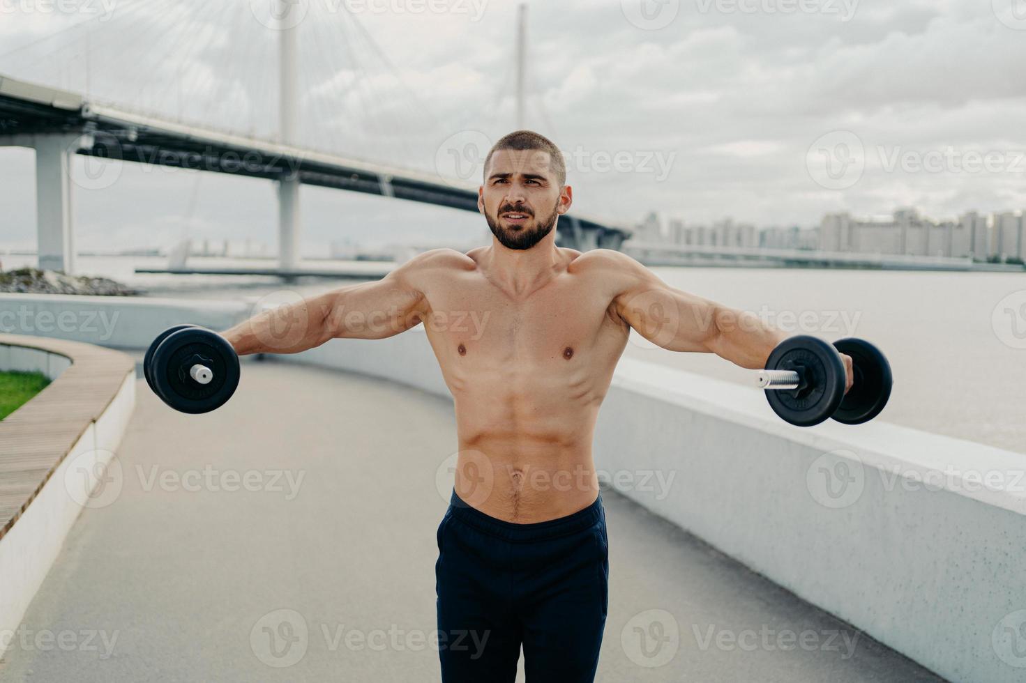 Motivated bodybuilder raises barbells outdoor enjoys great training trains muscles concentrated somewhere poses with naked muscular torso. Athletic shirtless sportsman has workout in open air photo