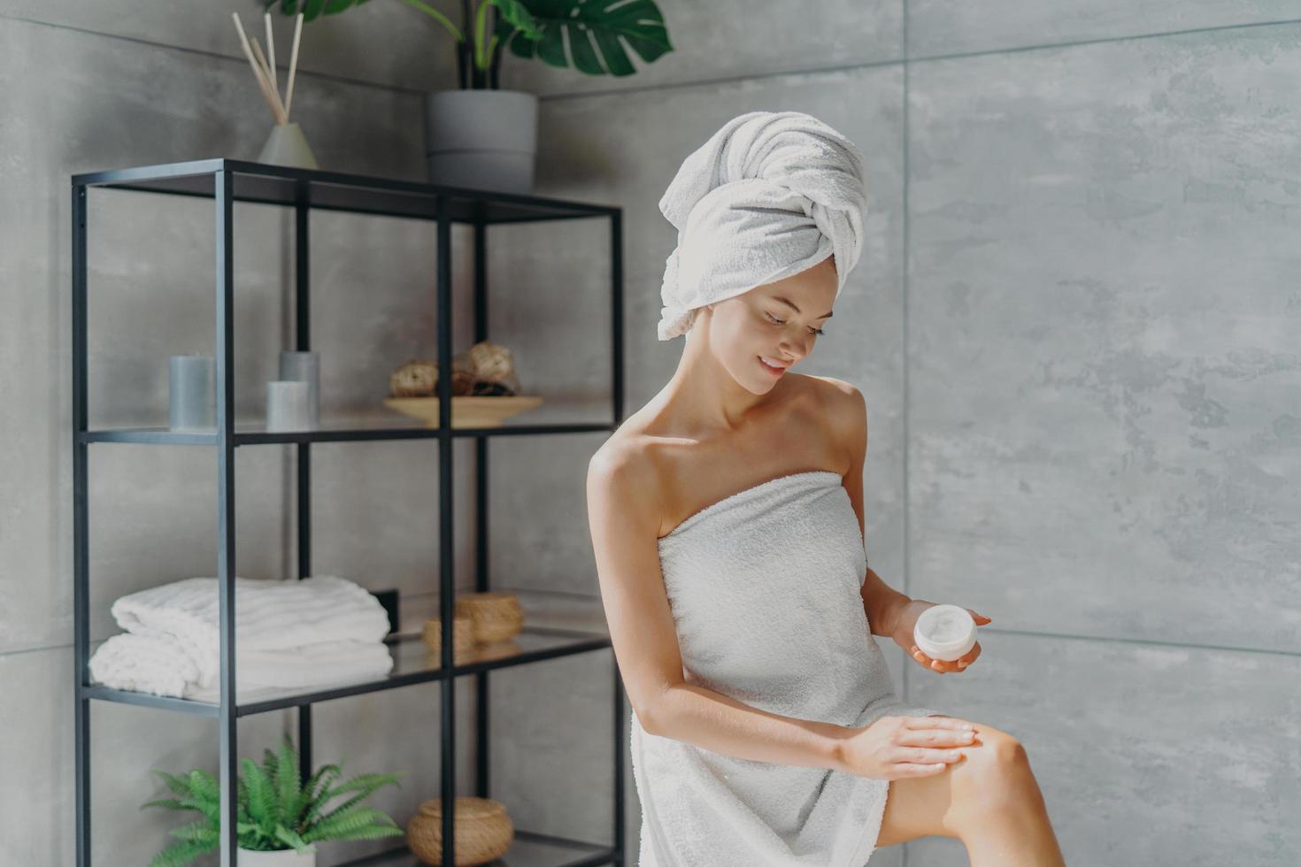 Body care concept. Lovely female model applies moisturising cream on leg, has healthy soft skin after taking bath, wrapped in towel, poses against bathroom interior. Beauty routine, wellness photo