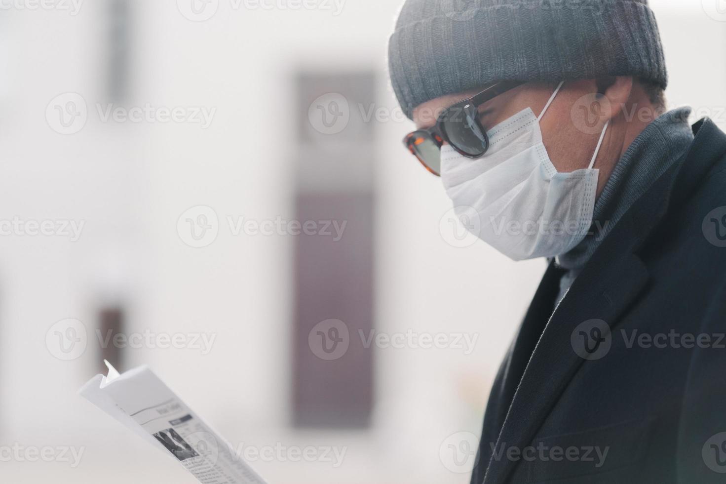 Sideways shot of man in hat, sunglasses and protective mask, reads newspaper outdoor, poses at street, finds out news from press during coronavirus outbreak and quarantine, respiratory virus photo