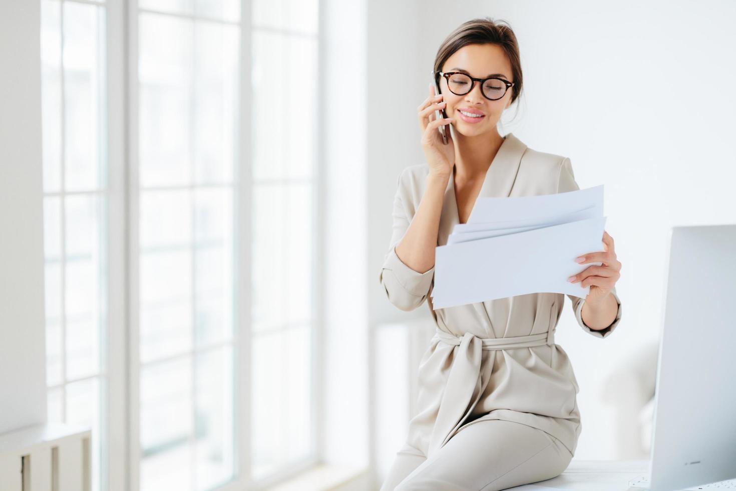 Attractive businesswoman reads papers or business documents, has telephone talk with business partner, examines paperwork before meeting, sits at desktop, smiles happily, works with documentation photo