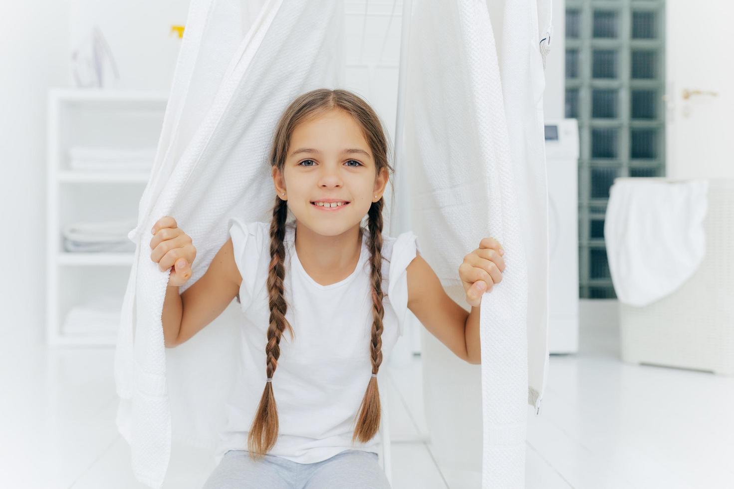 feliz chica europea con dos trenzas, posa cerca del tendedero entre sábanas  blancas secas, posa en la sala de lavado contra un fondo borroso, color  blanco. niños, limpieza, concepto de lavado. 10359420