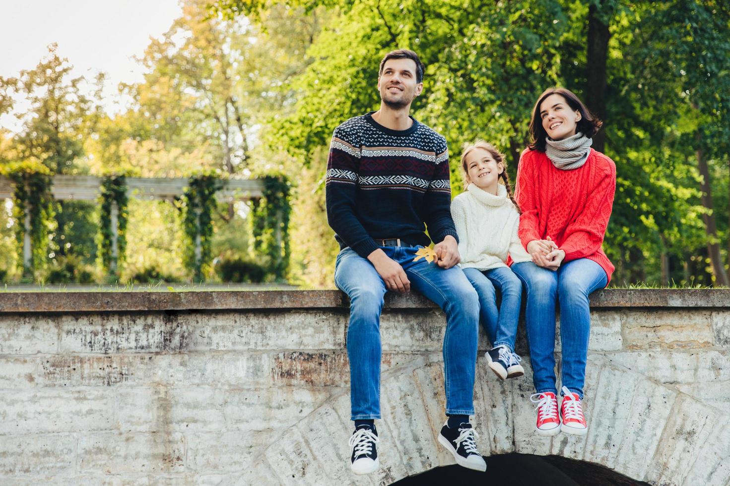 Horizontal portrait of adorable child sits between her mother and father, enjoy beaitiful landscapes from top of ancient bridge, breath fresh air, look into distance. Family and relationship concept photo