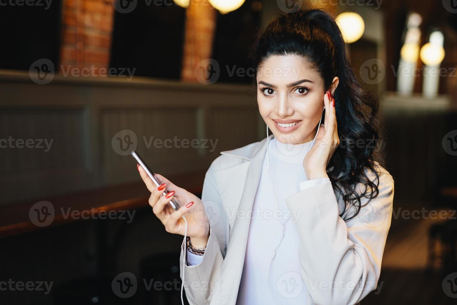 Cute woman with dark pony tail having long eyelashes and expressive eyes sitting at cafe listening to music online enjoying high-speed internet connection. People, beauty and technology concept photo
