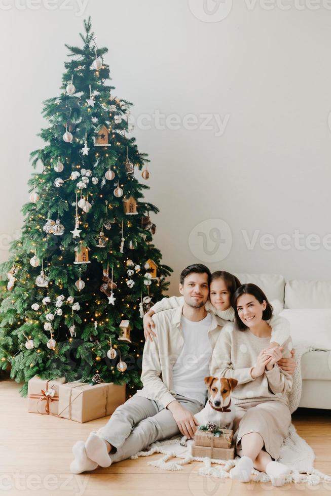 Three family members pose on floor in cozy room, gift boxes around, decorated New Year tree and sofa. Father, mother and daughter with jack russell terrier dog anticipate for coming holiday. photo