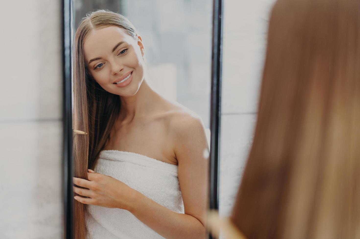foto de una mujer encantadora que se cepilla el cabello largo y saludable con un peine, se mira en el espejo, cuida su belleza, se envuelve en una toalla de baño, posa en el baño. cuidado del cabello y concepto de belleza para mujeres