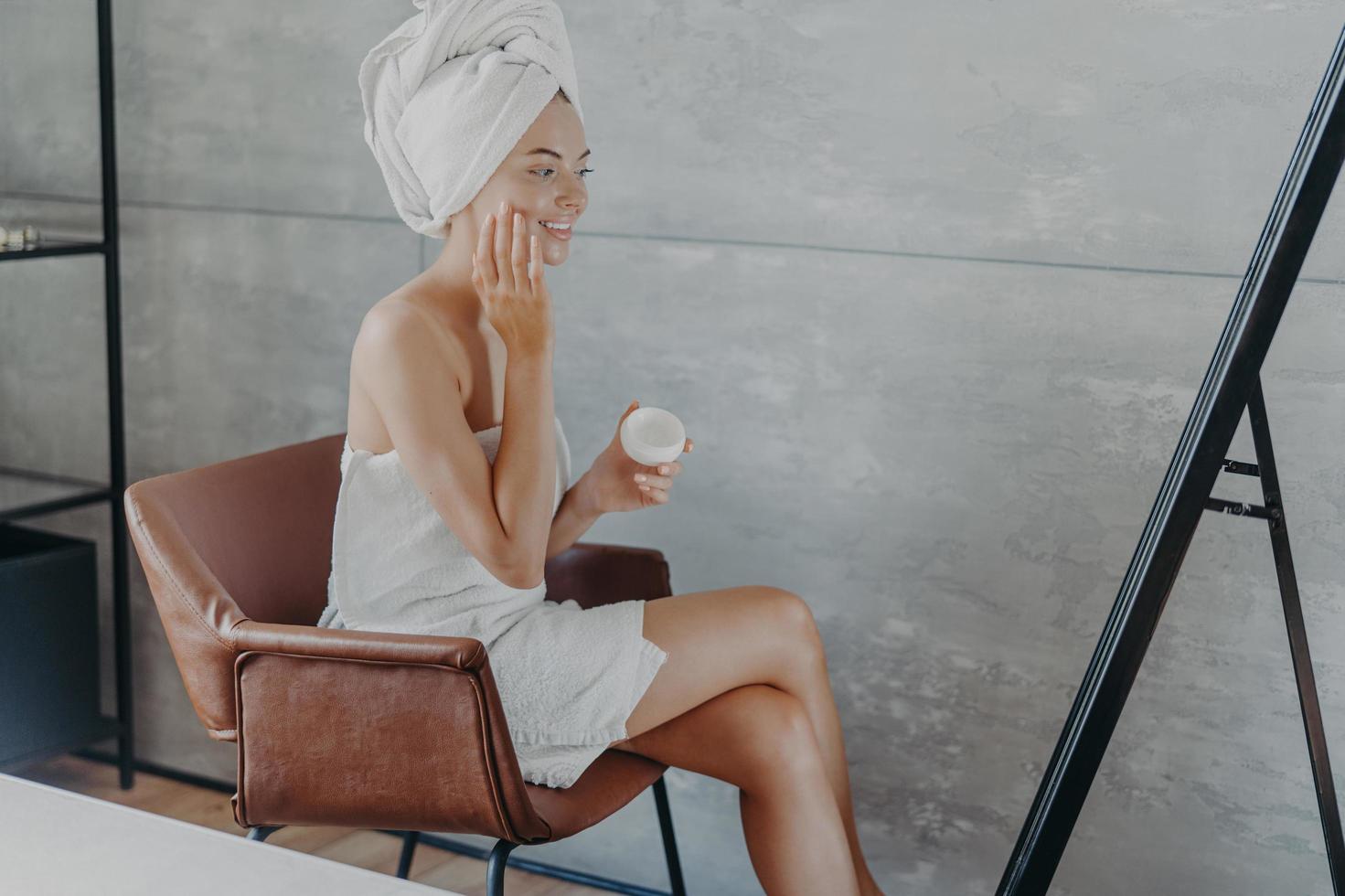 Skin care concept. Indoor shot of pretty smiling woman sits in comfortable armchair opposite mirror, applies cosmetic cream on face, wears wrapped towel on head, wants to have well groomed complexion photo