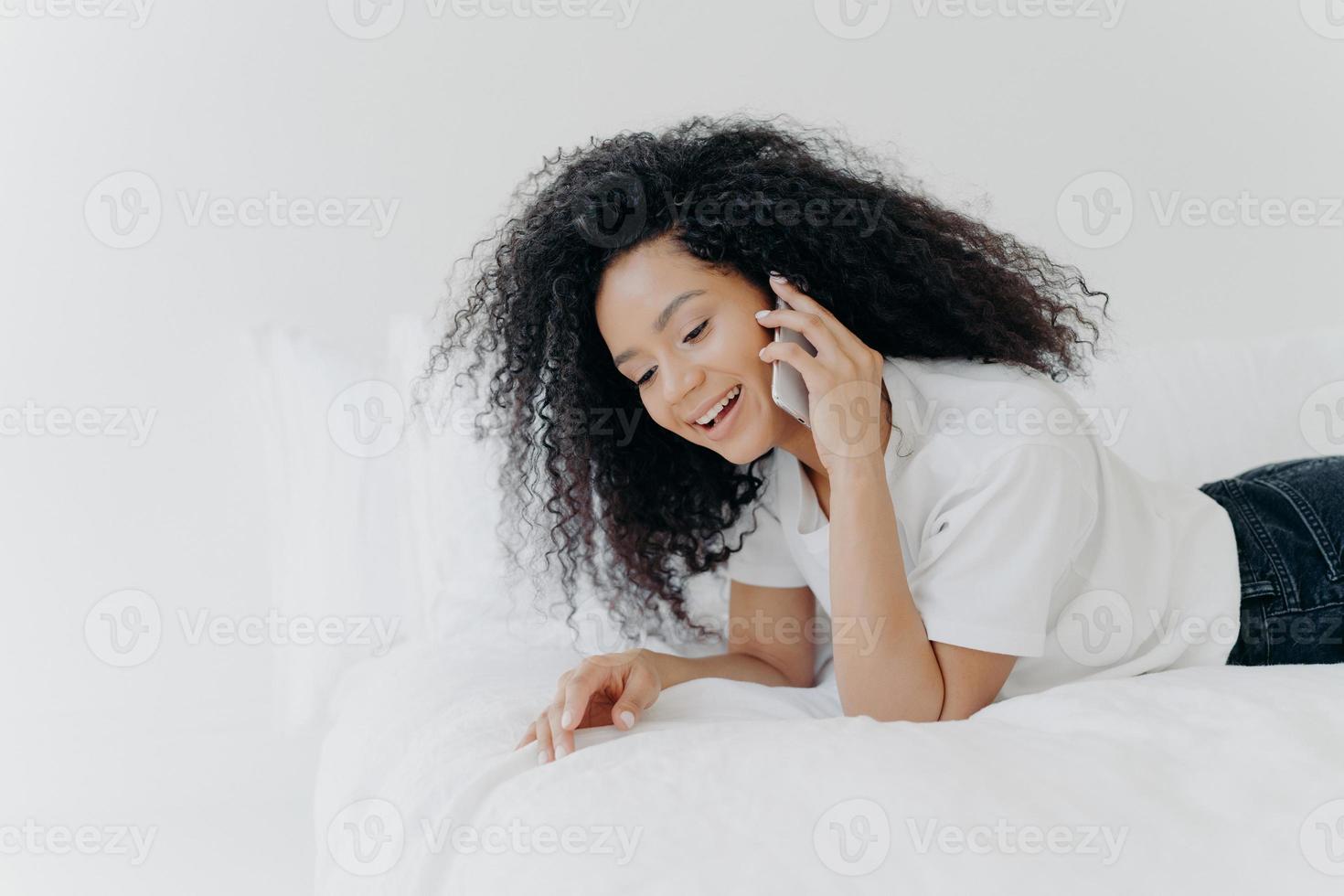 una mujer relajada y complacida con el pelo rizado tiene una conversación telefónica agradable, habla con un amigo durante el fin de semana antes de dormir, usa una camiseta blanca, se acuesta en una cama acogedora, mantiene un aparato moderno cerca de la oreja foto