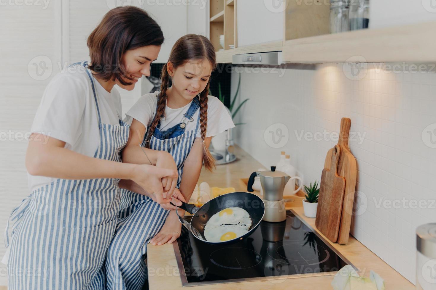 Busy mother and daughter wear striped aprons, pose at kitchen near cooker, fry eggs on pan, prepare fast breakfast, enjoy domestic atmosphere. Mum teaches small kid to cook. Happy family concept photo