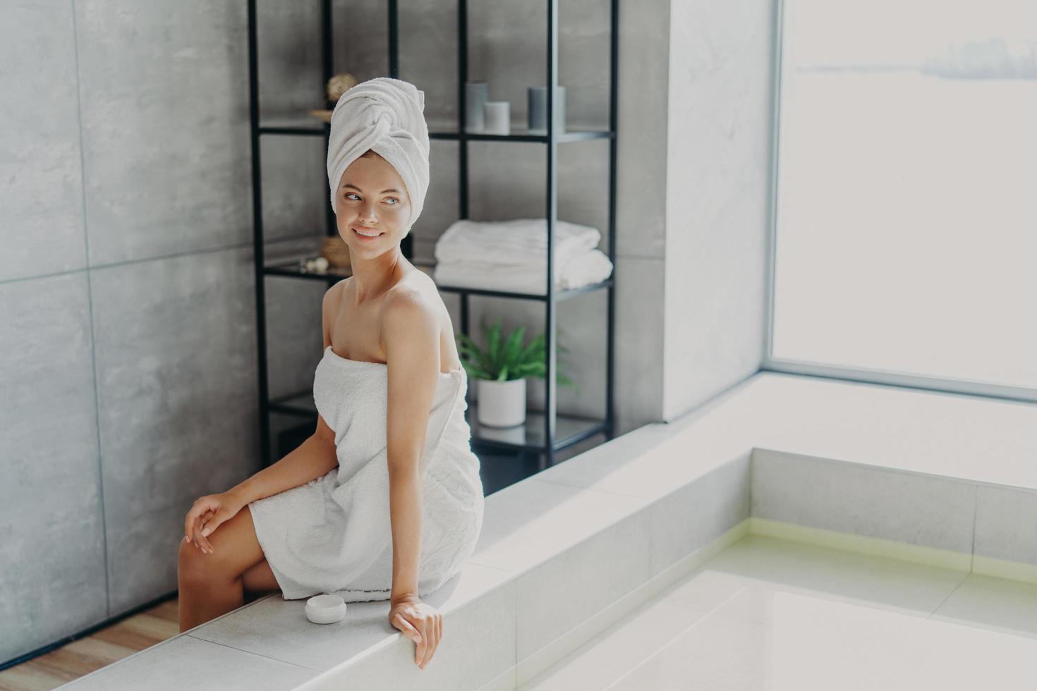 Beauty model with healthy perfect skin applies cream for body care, poses wrapped in towel after taking shower, poses at bathtube, looks thoughtfully away, feels refreshed and relaxed. Spa procedures photo