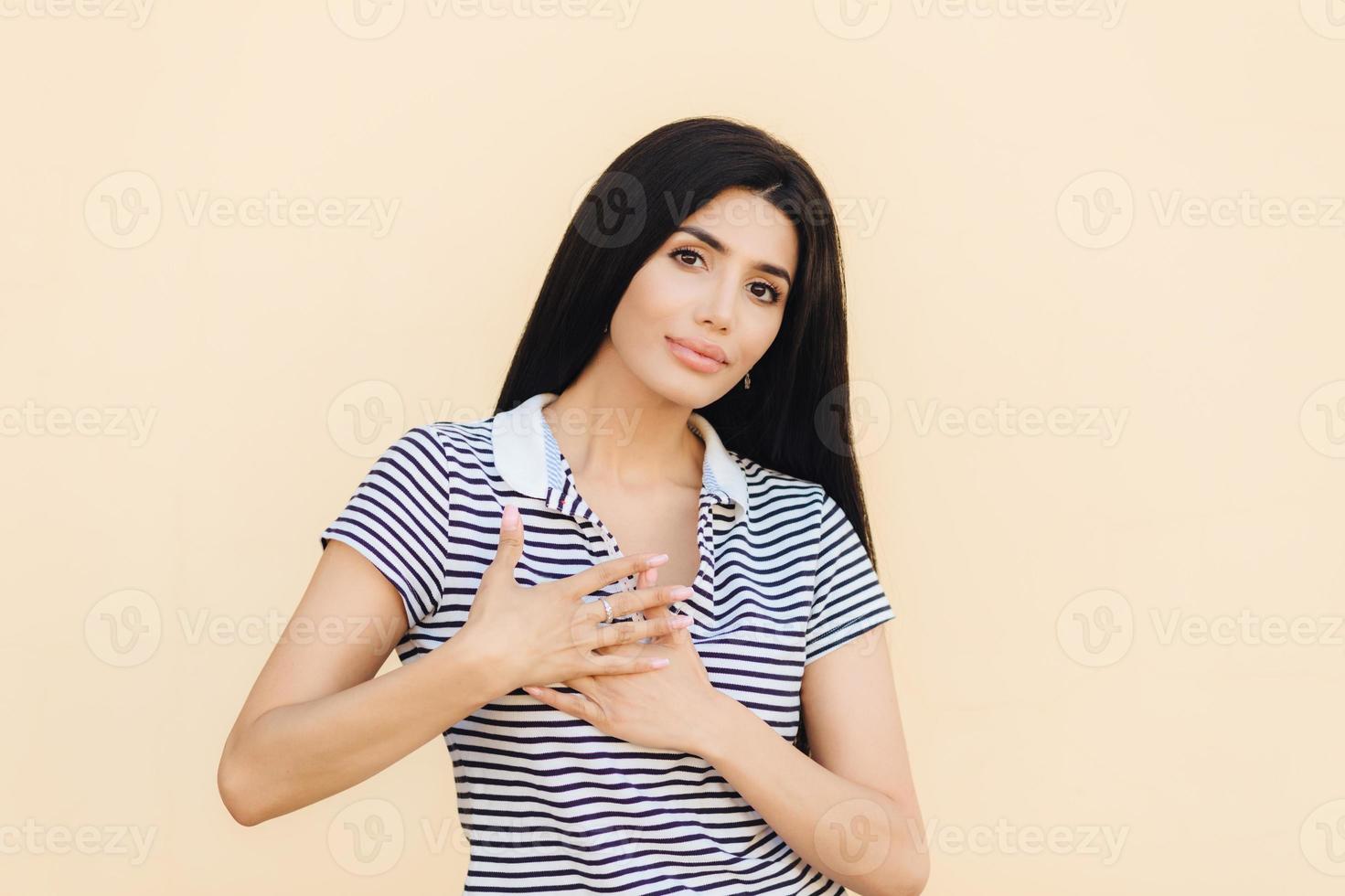 Friendly looking brunette female keeps hands on heart as expresses her gratitude, has serious expression, wears casual clothes, isolated over studio background. People, body language concept photo