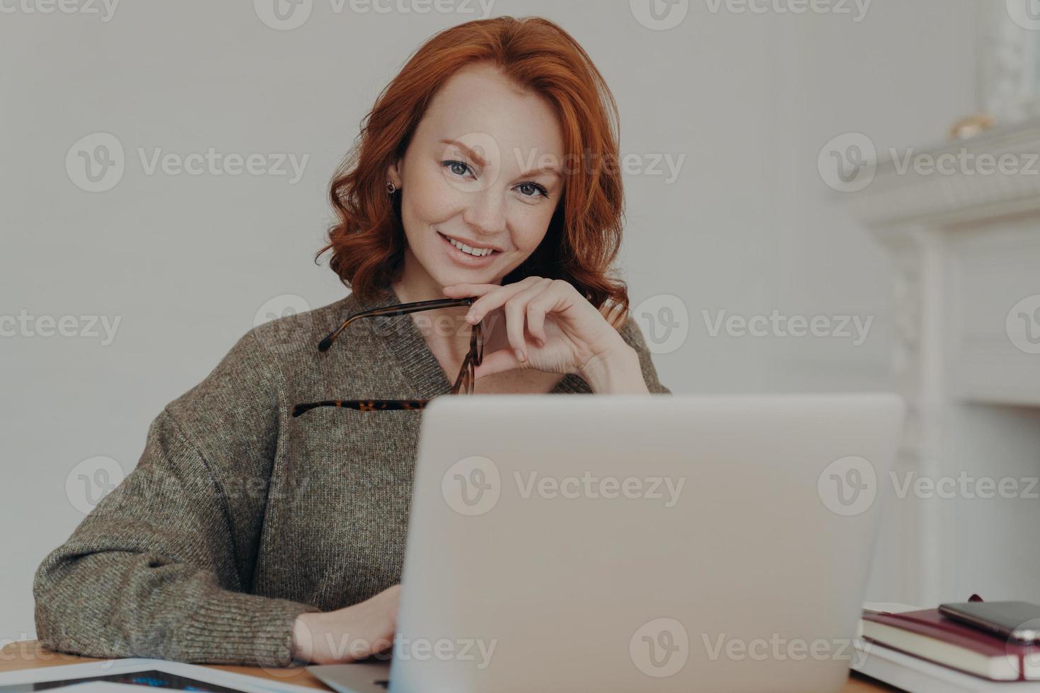 Successful professional businesswoman with red hair works remotely on laptop computer, reads article about business affairs online, sits at desktop, uses modern technologies, prepares financial report photo