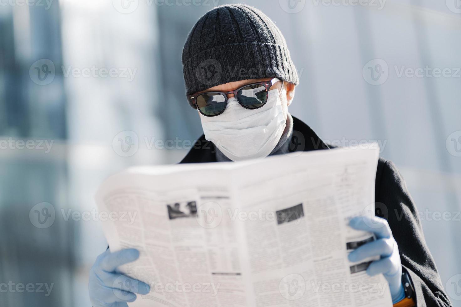 Photo of European man wears protective medical mask and gloves, walks through city outdoor, reads article about virus outbreak in different countries uses protective measures during coronavirus spread