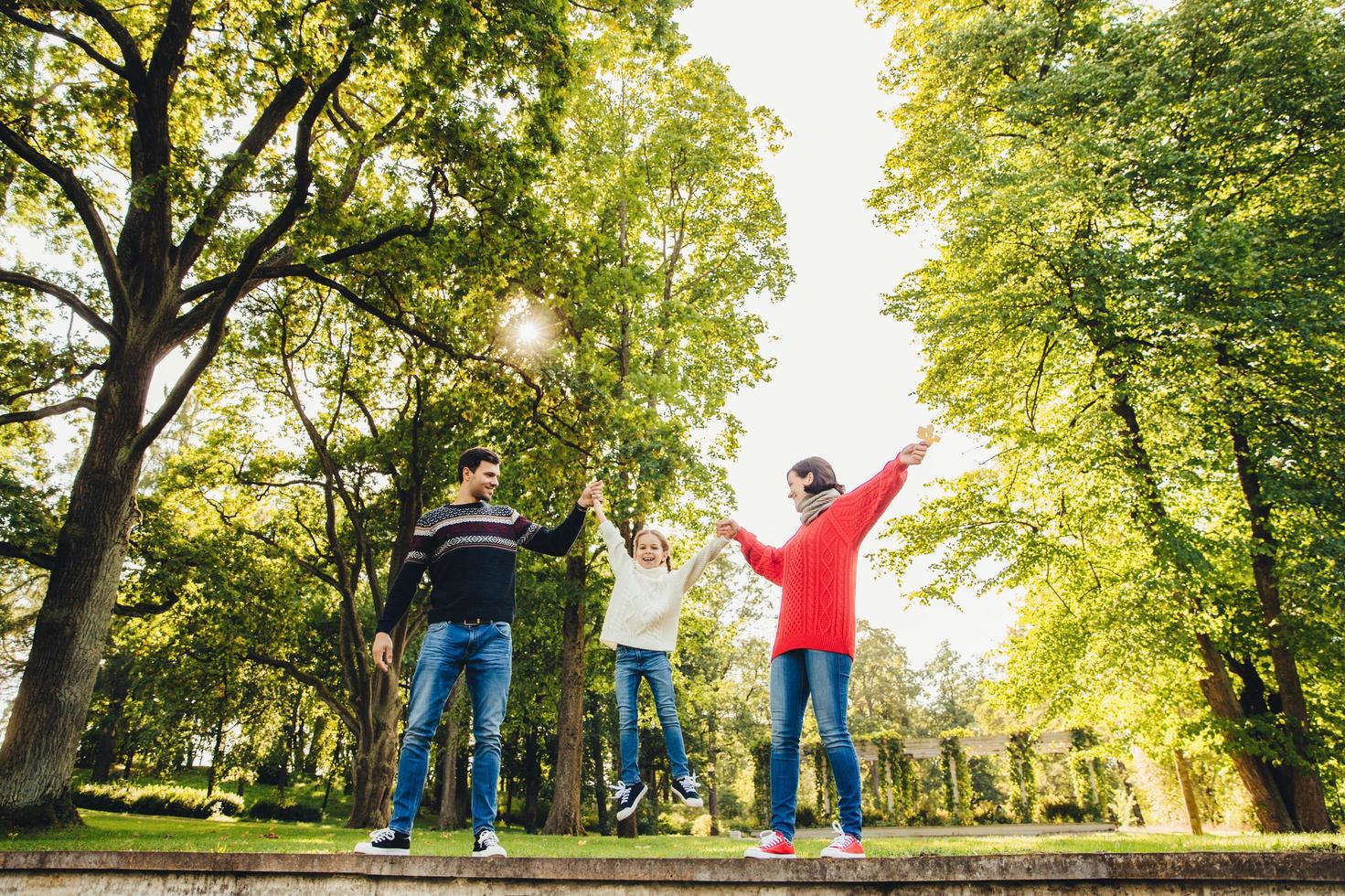 Portrait of friendly affectionate family have fun together, spend free time outdoors, stand against green trees in park, try to rock small child in hands. Three family memebers play on nature photo