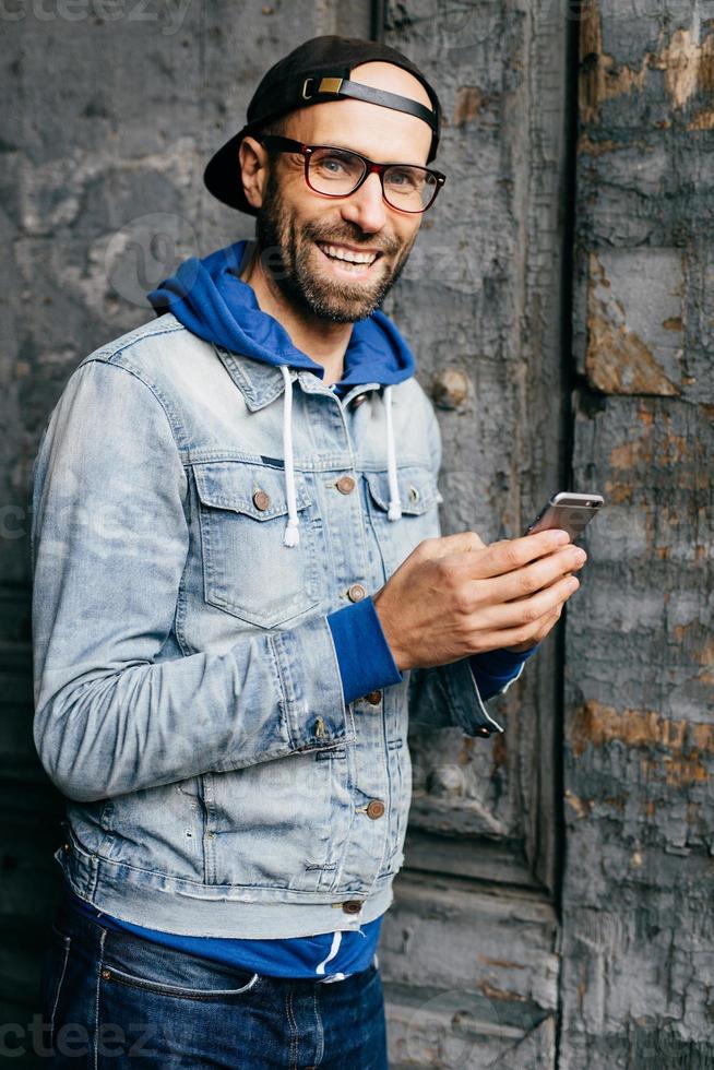 Stunning man with blue eyes, beard and broad smile wearing cap, denim shirt and eyeglasses holding smartphone isolated over old wall background. Pleased man in stylish clothes using cell phone photo