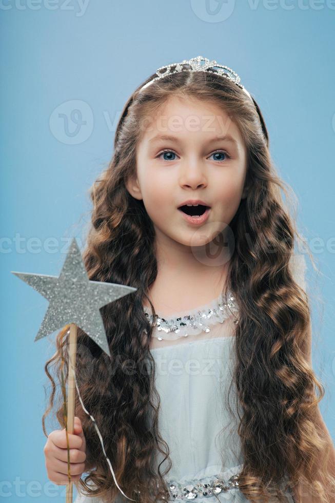 Small little child female princess wears crown and dress, holds magic wand, has long dark curly hair poses over blue background. Beautiful kid preapres for carnival or festive event. Childhood concept photo