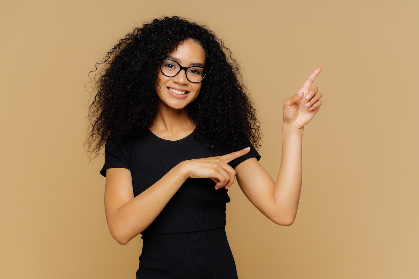 Isolated shot of happy African American woman points up and aside, demonstrates blank copy space for your advertisement, wears spectacles, casual t shirt, has gentle smile on face. People and promo photo