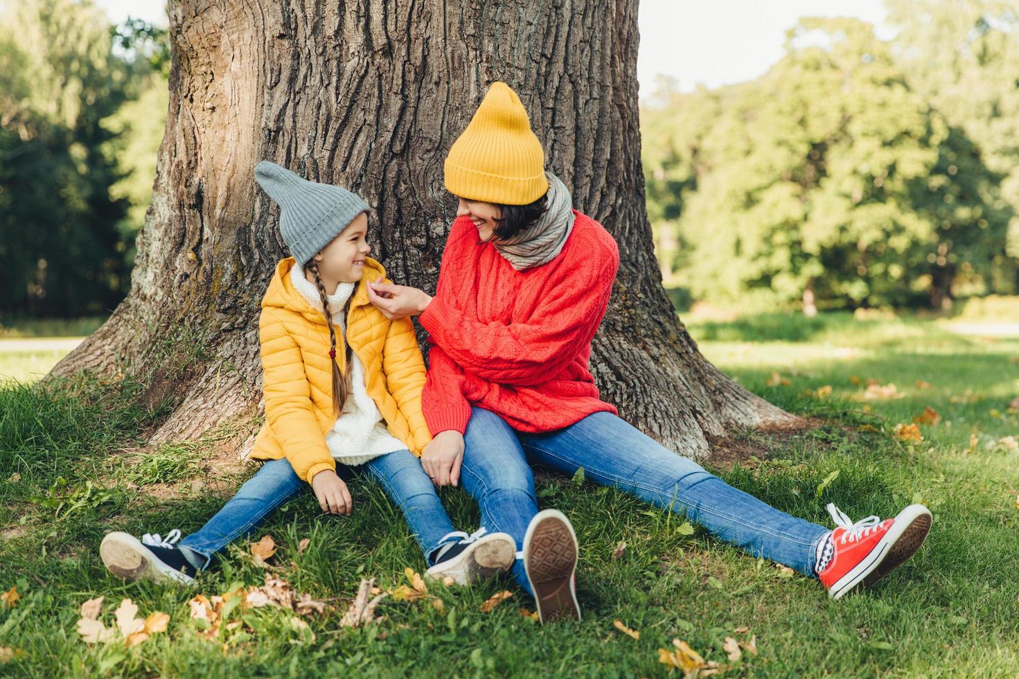 Little daughter and her mother have fun together, dressed warm, sit near big tree on green grass, look at each other with love. Affectionate mom and small female kid have good mood, spend weekends photo