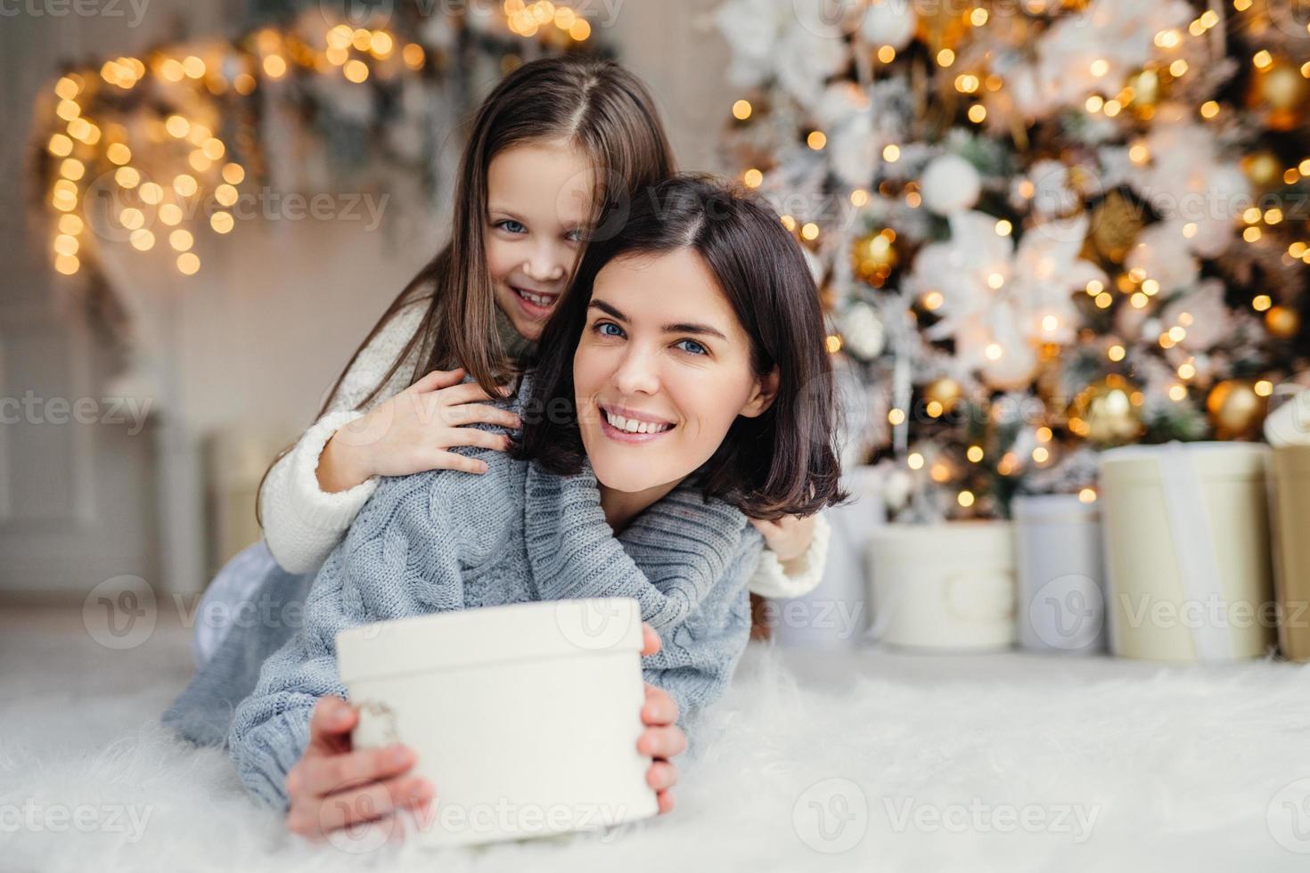 This present is for you  Happy small kid embraces her affectionate mother who holds wrapped present, stands against decorated background with garlands and New Year tree. Domestic atmosphere. photo