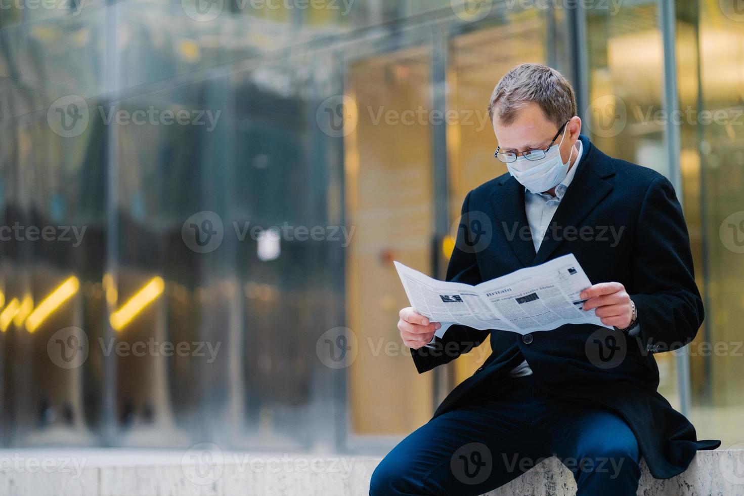 Outdoor shot of serious man boss takes break after walking, reads newspaper, wears spectacles for good vision, medical mask to protect himself from viruses, finds out news about spreading coronavirus photo