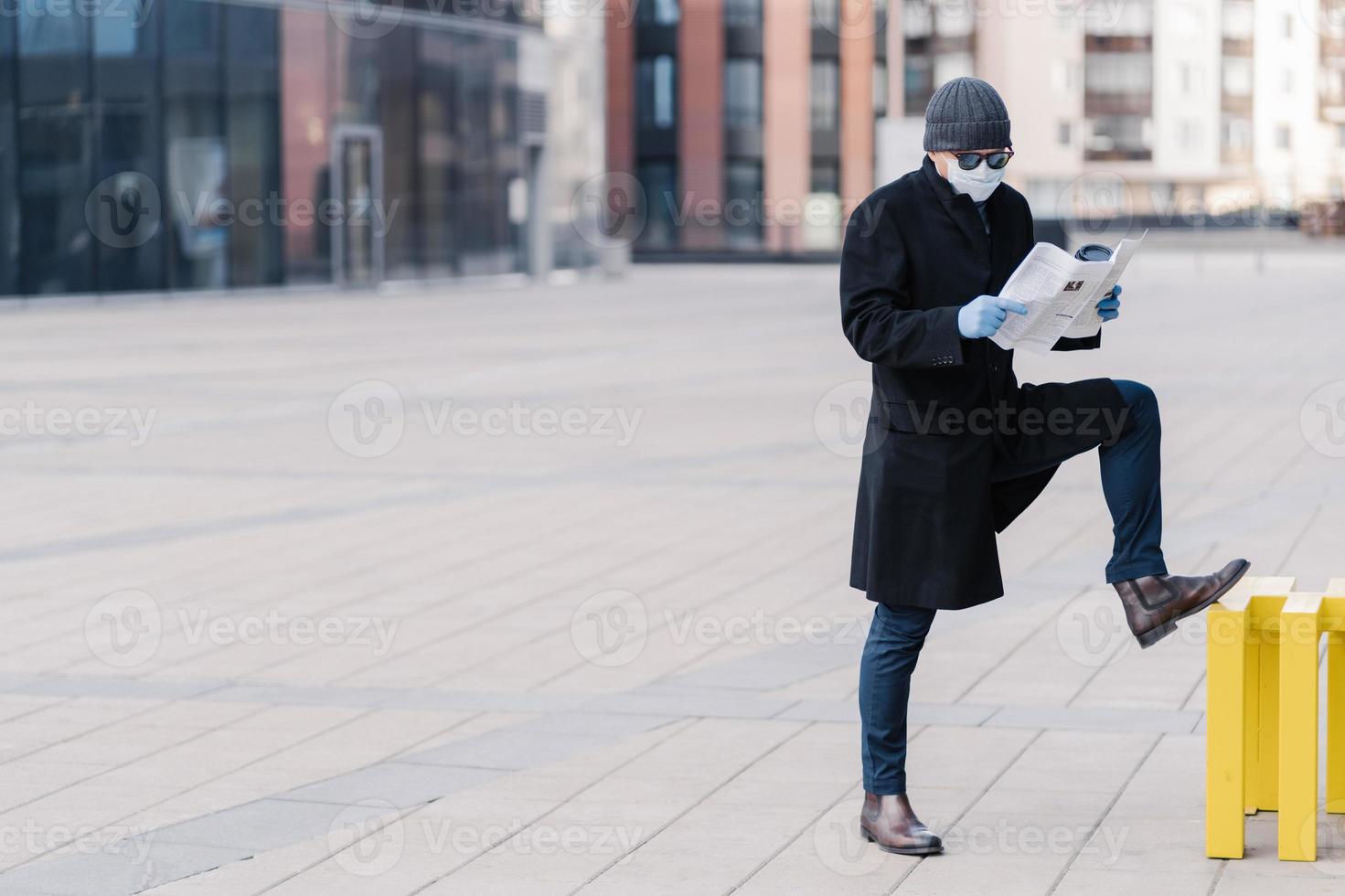 Sick man with flu wears medical mask to protect spreading infectious disease, dressed in coat, stands at street near office building, reads newspaper, finds out news about coronavirus pandemic photo