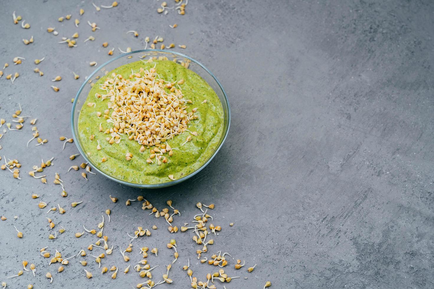 Horizontal shot of glass bowl with fresh vegetetable green smoothie containig much vitamins, buckwheat sprouts around and on top. Dark background. Copy space photo