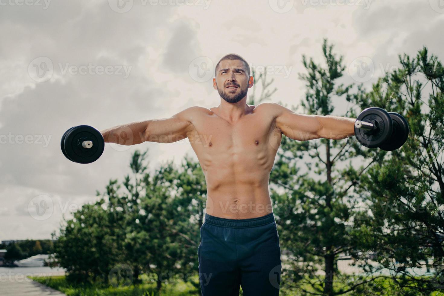 Strong handsome muscular bodybuilder pumps up arm muscles with barbells, does bidybuilding biceps exercises outdoor, poses with naked torso against green trees and sky, makes weight exercise photo