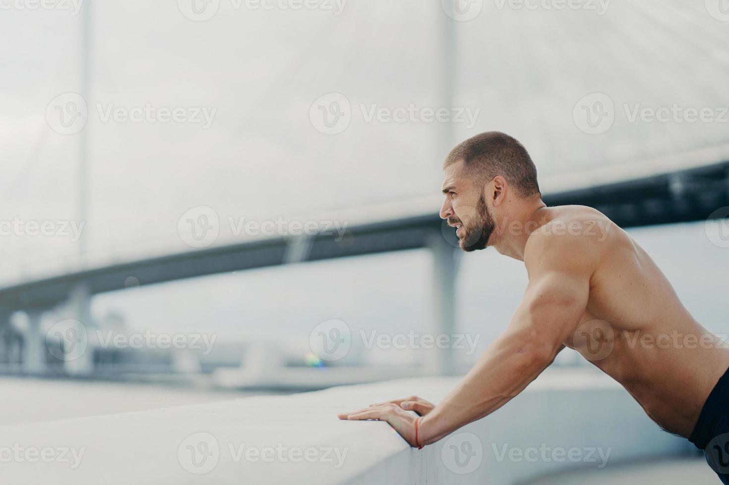 Motivated male sportsman does push up exercise poses outdoor at bridge concentrated into distance has morning workout naked muscular torso warms up before jogging. Healthy lifestyle concept. photo