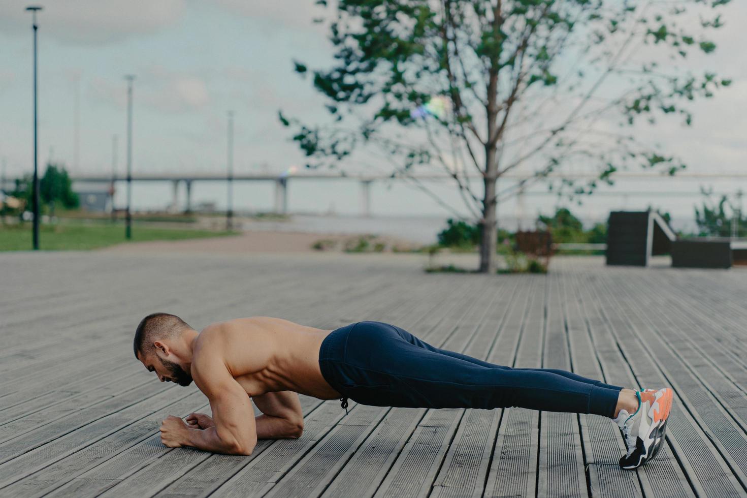 Horizontal shot of sporty bearded man stands in plank pose, practices yoga outdoor and dressed in active wear, breathes fresh air. Sport, fitness and healthy lifestyle concept. Training workout photo