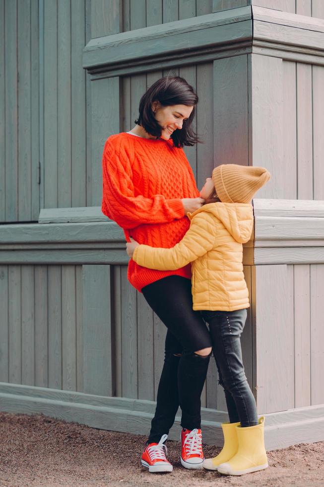 toma vertical de una madre atractiva y cariñosa mira a los ojos de su hija con amor, abraza mientras posa afuera, usa jeans rasgados, suéter y zapatillas, sonríe positivamente. concepto de relación familiar foto