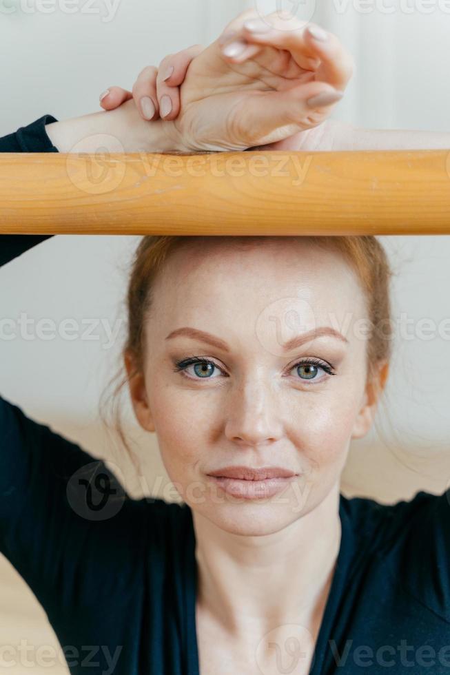 Headshot of talented skilled female ballet dancer poses near barre, has blue eyes, healthy skin, appealing appearance, practices dance in dancing hall. Red haired choreography instructor indoor photo