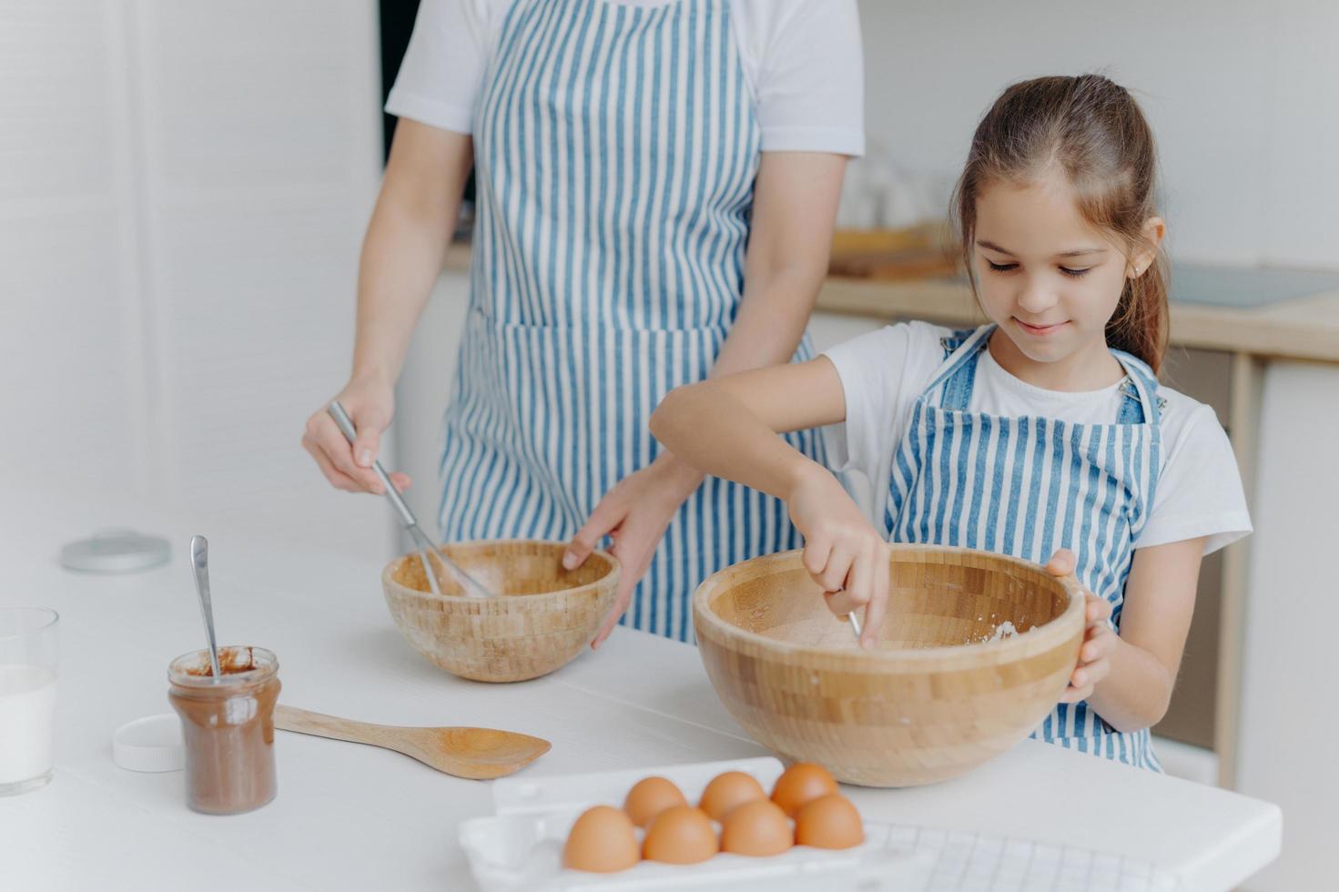 Mother gives culinary lesson to little child, stand next to each other, mix ingredient in big wooden bowls, make dough together, use eggs, flour and other products. Family, cooking, motherhood photo