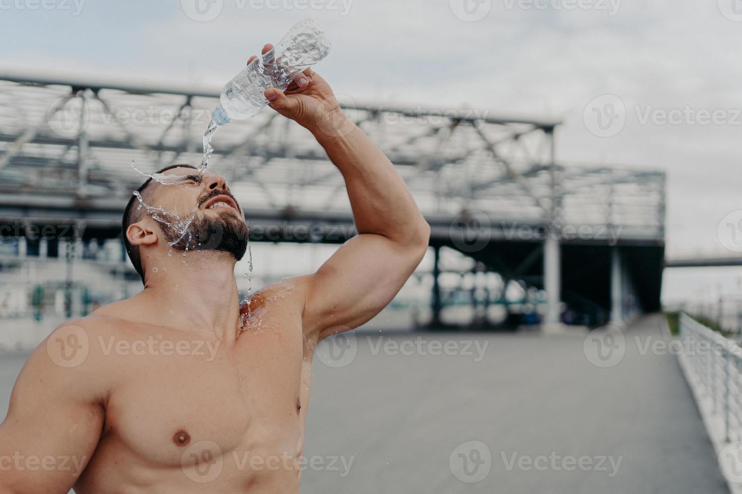 Tired sportsman cools off with fresh water poses with naked torso outdoor, tries to refresh himself, leads active lifestyle, takes break after cardio training. Excercising, refreshment concept photo